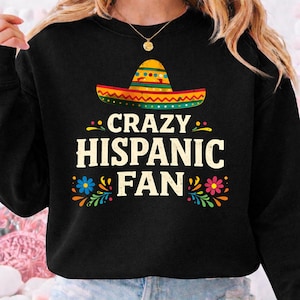 May include: Black sweatshirt with the text "CRAZY HISPANIC FAN" in white, featuring a colorful sombrero graphic. The design includes floral accents in blue, pink, and yellow.