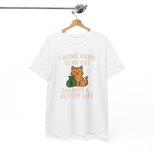 Könnte beinhalten: Weißes T-Shirt mit dem Text "I WORK HARD SO MY CAT CAN HAVE A BETTER LIFE". Die Grafik zeigt eine lächelnde orange Katze, die einen grünen Geldsack mit einem Dollarzeichen hält.