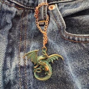 May include: A copper-colored chain with a dragon pendant attached to a denim jean. The dragon is green and gold with outstretched wings. The chain is attached to the jean's button.