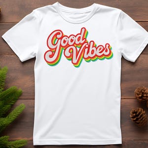 May include: A white t-shirt featuring the phrase "Good Vibes" in a retro style. The text is in red, yellow, green, and blue, with a shadow effect. The shirt has a crew neck and short sleeves.