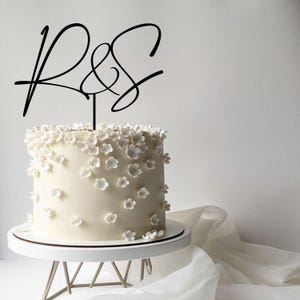 Peut inclure: Un gâteau blanc rond orné de fleurs blanches et surmonté d'un cake topper noir avec les lettres "R & S" en cursive. Le gâteau est posé sur un présentoir blanc avec une base en métal argenté.