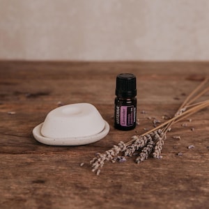 May include: A small, white ceramic essential oil diffuser with a matching dish, a bottle of lavender essential oil, and dried lavender sprigs on a wooden surface. The oil bottle has a black cap and a purple label.