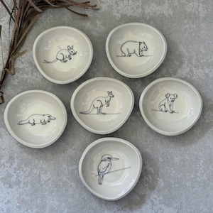 May include: Set of six small, off-white ceramic bowls. Each bowl features a black line drawing of an Australian animal: a rat, wombat, koala, kangaroo, platypus, and a bird. The bowls have an irregular, organic shape.