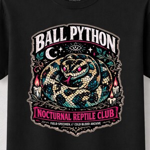 May include: A black short-sleeve t-shirt featuring a graphic of a coiled ball python with pink eyes. The design includes the text BALL PYTHON, NOCTURNAL REPTILE CLUB, and FIELD SPECIMEN // COLD BLOOD ARCHIVE, surrounded by candles, leaves, and a crescent moon.