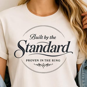 Può includere: T-shirt color crema con la scritta "Built by the Standard" in un design circolare. La parola "Standard" è in grassetto, con un carattere corsivo, con "Proven in the Ring" sotto. La maglietta è realizzata in materiale morbido.