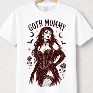 May include: A white short-sleeve t-shirt featuring a dark brown graphic illustration of a person in a corset and gloves with the text GOTH MOMMY. The design includes roses, crescent moons, stars, and bats surrounding the central figure.