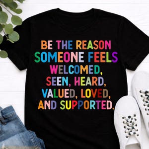 May include: Black t-shirt with the text "BE THE REASON SOMEONE FEELS WELCOMED, SEEN, HEARD, VALUED, LOVED, AND SUPPORTED" in colorful letters. The shirt is paired with blue jeans and white sneakers. The words "BLACK premium cotton" are also visible.