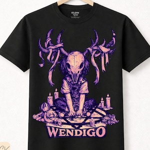 Puede incluir: Camiseta negra con un diseño Wendigo morado y rosa. El gráfico muestra una calavera con cuernos, velas y la palabra "WENDIGO". Hecha de algodón premium.