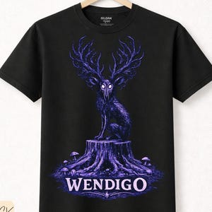 Puede incluir: Camiseta negra con un diseño morado de Wendigo. El gráfico muestra una criatura con cuernos y una cara de calavera, sentada en un tocón de árbol. La palabra "WENDIGO" está impresa debajo. Hecha de algodón premium.