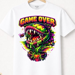 Könnte beinhalten: Weißes T-Shirt mit einer farbenfrohen Grafik einer fleischfressenden Pflanze und dem Schriftzug "GAME OVER" in einer leuchtenden, mehrfarbigen Schriftart. Das T-Shirt ist aus Premium-Baumwolle.