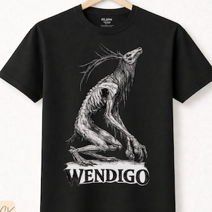 Puede incluir: Camiseta negra con una ilustración detallada de una criatura esquelética con cuernos, etiquetada como "WENDIGO" en texto blanco. La camiseta está hecha de algodón de primera calidad y está colgada en una percha de madera.