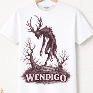 Puede incluir: Camiseta blanca con una ilustración oscura de una criatura con cuernos y la palabra "WENDIGO". El diseño incluye árboles desnudos y un suelo texturizado. La camiseta está hecha de algodón de primera calidad.