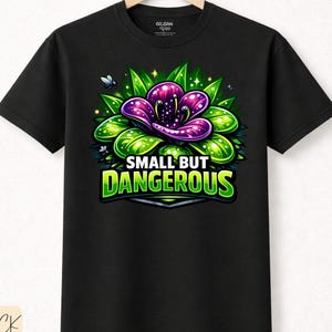 Könnte beinhalten: Schwarzes T-Shirt mit einem farbenfrohen Grafikdesign einer lila und grünen Blume und dem Text "SMALL BUT DANGEROUS". Das T-Shirt besteht aus hochwertiger Baumwolle und hängt an einem Holzbügel. Das Design beinhaltet kleine fliegende Insekten.