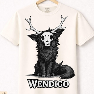 Puede incluir: Camiseta color crema con una ilustración negra de una criatura con cuernos y una cara de calavera. La palabra "WENDIGO" está impresa en negrita, en negro, debajo de la ilustración. Hecha de algodón natural de primera calidad.