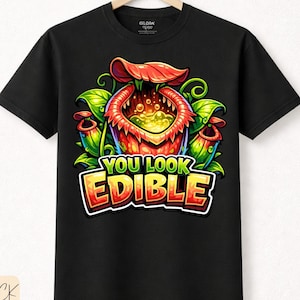 Könnte beinhalten: Schwarzes T-Shirt mit einer farbenfrohen Cartoon-Illustration einer fleischfressenden Pflanze und dem Text "YOU LOOK EDIBLE" in einer verspielten Schriftart. Aus hochwertiger Baumwolle.