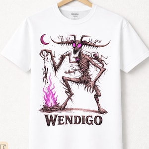 Puede incluir: Camiseta blanca con una ilustración detallada de una criatura esquelética con cuernos, una luna creciente y una llama púrpura. La palabra "WENDIGO" está impresa en negrita debajo del gráfico. La camiseta es de algodón premium.
