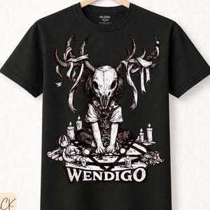Puede incluir: Camiseta negra con una ilustración detallada de una criatura con rostro de calavera y cornamenta, rodeada de velas y otros objetos. La palabra "WENDIGO" está impresa en blanco. La camiseta es de algodón premium.