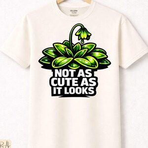 Könnte beinhalten: Cremefarbenes T-Shirt mit einer leuchtend grünen Pflanzen-Grafik und dem Text "NOT AS CUTE AS IT LOOKS" in Schwarz und Weiß. Das Shirt besteht aus natürlicher Premium-Baumwolle und wird auf einem Holzbügel präsentiert.