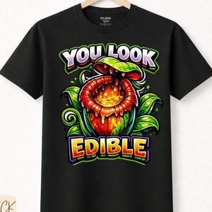 Könnte beinhalten: Schwarzes T-Shirt mit einem farbenfrohen Grafikdesign einer fleischfressenden Pflanze und dem Text "YOU LOOK EDIBLE". Das Design verwendet leuchtende Farben wie Grün, Rot und Gelb, was einen auffälligen Look erzeugt.