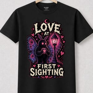 May include: A black t-shirt featuring a graphic of a fuzzy moth-like creature with purple wings standing next to a streetlamp. The creature holds a pink heart. The text reads LOVE AT FIRST SIGHTING in a white, distressed, gothic-style font.
