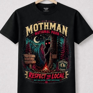 Può includere: T-shirt nera con un design grafico colorato per il Mothman National Park. Il design include il testo "Mothman National Park" e "Respect the Local" con una scena forestale, una creatura e una luna crescente.