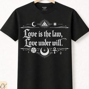 May include: Black t-shirt featuring the white text "Love is the law, Love under will." with occult symbols. Made from premium cotton. The t-shirt is hanging on a wooden hanger.