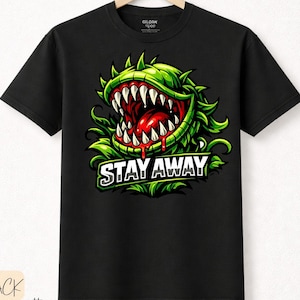 Könnte beinhalten: Schwarzes T-Shirt mit einem grünen Cartoon-Pflanzendesign, scharfen Zähnen und den Worten "STAY AWAY" in Weiß. Die Pflanze hat einen roten Mund und grüne Blätter. Das T-Shirt hängt an einem Holzbügel.