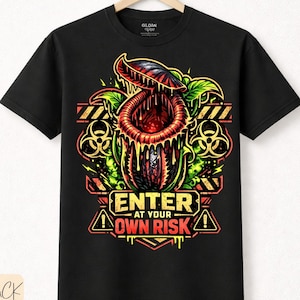 Könnte beinhalten: Schwarzes T-Shirt mit einem farbenfrohen Grafikdesign einer fleischfressenden Pflanze und dem Text "ENTER AT YOUR OWN RISK". Das Design beinhaltet Gefahrensymbole und Streifen. Aus hochwertiger Baumwolle.