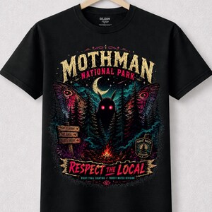 Può includere: T-shirt nera con grafica del Mothman National Park. Il design presenta una creatura simile a una falena con occhi rossi, una luna crescente e immagini della foresta. Il testo include "Respect the Local" e "Forest Watch Division".
