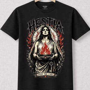 Könnte beinhalten: Schwarzes T-Shirt mit einer Grafik von Hestia, einer Frau, die eine Feuerschale hält, mit dem Text "Hestia" darüber und "Keep the Fire" darunter. Das Design umfasst rote und weiße Akzente, einen Schlüssel und Lorbeerblätter.