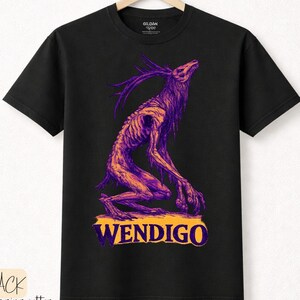 Puede incluir: Camiseta negra con una ilustración morada y naranja de un Wendigo. El diseño muestra una criatura esquelética con cuernos, arrodillada con la cabeza levantada. La palabra "WENDIGO" está impresa en naranja y morado.