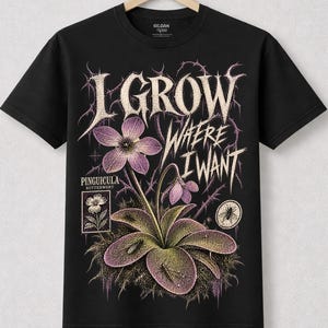May include: A black short-sleeve t-shirt featuring a graphic of a Pinguicula butterwort plant with purple flowers and green leaves. The text reads I GROW WHERE I WANT and PINGUICULA BUTTERWORT. Small insects are visible on the plant leaves.