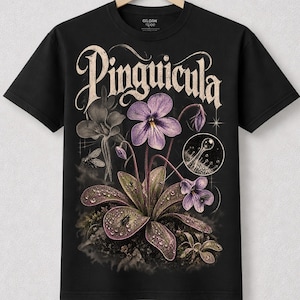 May include: A black short-sleeve t-shirt featuring a graphic print of a Pinguicula plant with purple flowers and dew-covered leaves. The design includes a small circular inset showing a magnified view of a leaf gland and the text Pinguicula.