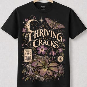 May include: A black short-sleeved t-shirt featuring a graphic print with the text "THRIVING IN THE CRACKS". The design includes a crescent moon, a moth, a Pinguicula plant, crystals, and small flowers in shades of purple, cream, and muted green.
