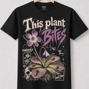 Peut inclure: Un t-shirt noir à manches courtes avec un motif de plante Pinguicula butterwort aux fleurs et feuilles violettes. Le texte indique This plant BITES, PINGUICULA BUTTERWORT, et SMALL PREY. BIG APPETITE. Un petit crâne est visible à la base.