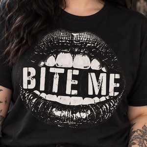 May include: A black short-sleeved t-shirt featuring a large white graphic print of a pair of lips with teeth and the text "BITE ME" centred on the chest. The design has a distressed, textured appearance against the solid black fabric.