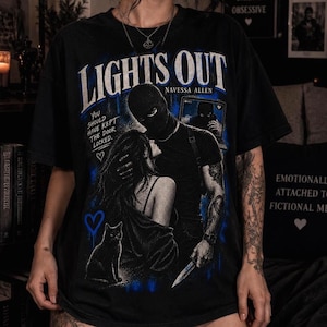 以下が含まれることがあります： LIGHTS OUTおよびNAVESSA ALLENのテキストがプリントされた黒の半袖Tシャツ。デザインには、目出し帽を被りナイフを持った人物、長い髪の人物、猫、そしてYOU SHOULD HAVE KEPT THE DOOR LOCKEDというテキストが描かれています。