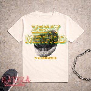 Può includere: T-shirt color crema con la scritta "ZESTY METHOD" in un design 3D sopra un'illustrazione in scala di grigi di una bocca sorridente. La frase "IS SO UNDERRATED" è sotto. Sono visibili una catena e una sneaker.