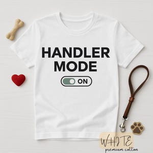 Può includere: T-shirt bianco con la scritta "HANDLER MODE ON" in nero. Il design include un interruttore a levetta. Sono visibili anche un osso per cani, un cuore rosso, un guinzaglio e un ciondolo a forma di zampa. La maglietta è in cotone premium.