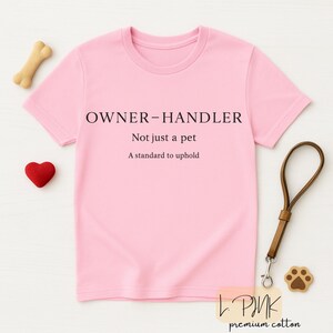May include: A pale pink t-shirt with the words "OWNER-HANDLER" and the phrases "Not just a pet, A standard to uphold." A dog bone, a red heart, a dog leash, and a paw-shaped charm are also visible.
