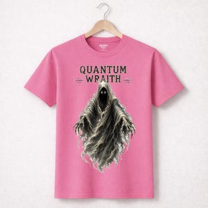 以下が含まれることがあります： 「QUANTUM WRAITH」の文字と、光る目とぼろぼろのマントを着たフード付きの人物のイラストが描かれたピンク色のTシャツ。Tシャツは木製のハンガーに掛けられています。デザインはファンタジーまたはホラーをテーマにしている可能性があります。