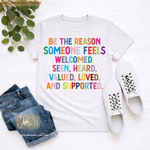 May include: A white t-shirt with the text "BE THE REASON SOMEONE FEELS WELCOMED, SEEN, HEARD, VALUED, LOVED, AND SUPPORTED" in colorful letters. The shirt is paired with blue denim jeans and white sneakers with black and white laces.