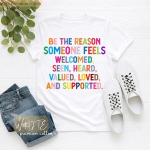 May include: White t-shirt with the text "BE THE REASON SOMEONE FEELS WELCOMED, SEEN, HEARD, VALUED, LOVED, AND SUPPORTED" in various colors. The shirt is paired with blue jeans and white sneakers. The words "WHITE premium cotton" are also visible.