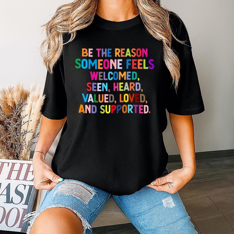 May include: Black t-shirt with the text "BE THE REASON SOMEONE FEELS WELCOMED, SEEN, HEARD, VALUED, LOVED, AND SUPPORTED" in rainbow colors. The shirt has a crew neck and short sleeves. The shirt is made of a soft material.