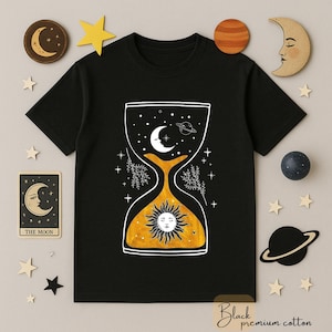 May include: Black t-shirt with a celestial hourglass design. The hourglass features a sun, moon, stars, and planets in white and yellow. The words "Black premium cotton" are at the bottom of the shirt.