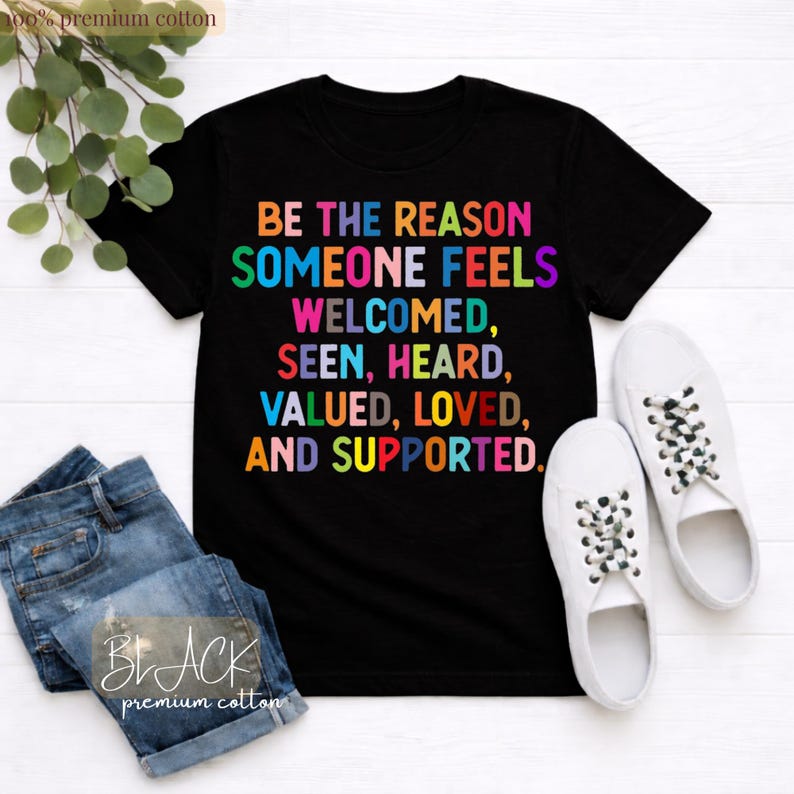 May include: Black t-shirt with the text "BE THE REASON SOMEONE FEELS WELCOMED, SEEN, HEARD, VALUED, LOVED, AND SUPPORTED" in colorful letters. The shirt is paired with blue jeans and white sneakers. The words "BLACK premium cotton" are also visible.