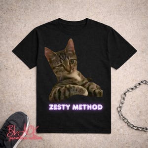 Può includere: T-shirt nero con grafica di gatto e la scritta "ZESTY METHOD". Il gatto è marrone e grigio con occhi verdi. Il testo è in un font in stile neon. La maglietta è a girocollo.
