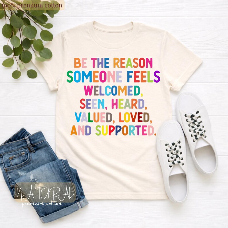 May include: Cream-colored t-shirt with the text "BE THE REASON SOMEONE FEELS WELCOMED, SEEN, HEARD, VALUED, LOVED, AND SUPPORTED" in various colors. The shirt is paired with blue denim shorts and white sneakers.