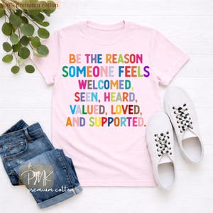 May include: A light pink t-shirt with the text "BE THE REASON SOMEONE FEELS WELCOMED, SEEN, HEARD, VALUED, LOVED, AND SUPPORTED" in colorful letters. The shirt is paired with blue jeans and white sneakers.