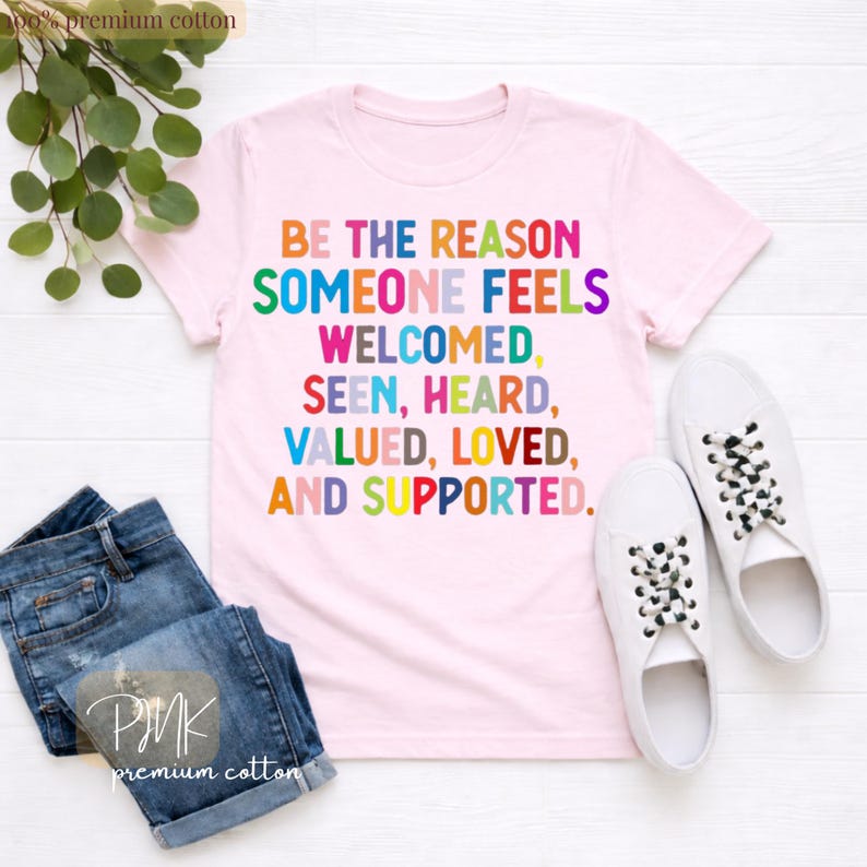 May include: A light pink t-shirt with the text "BE THE REASON SOMEONE FEELS WELCOMED, SEEN, HEARD, VALUED, LOVED, AND SUPPORTED" in colorful letters. The shirt is paired with blue jeans and white sneakers.